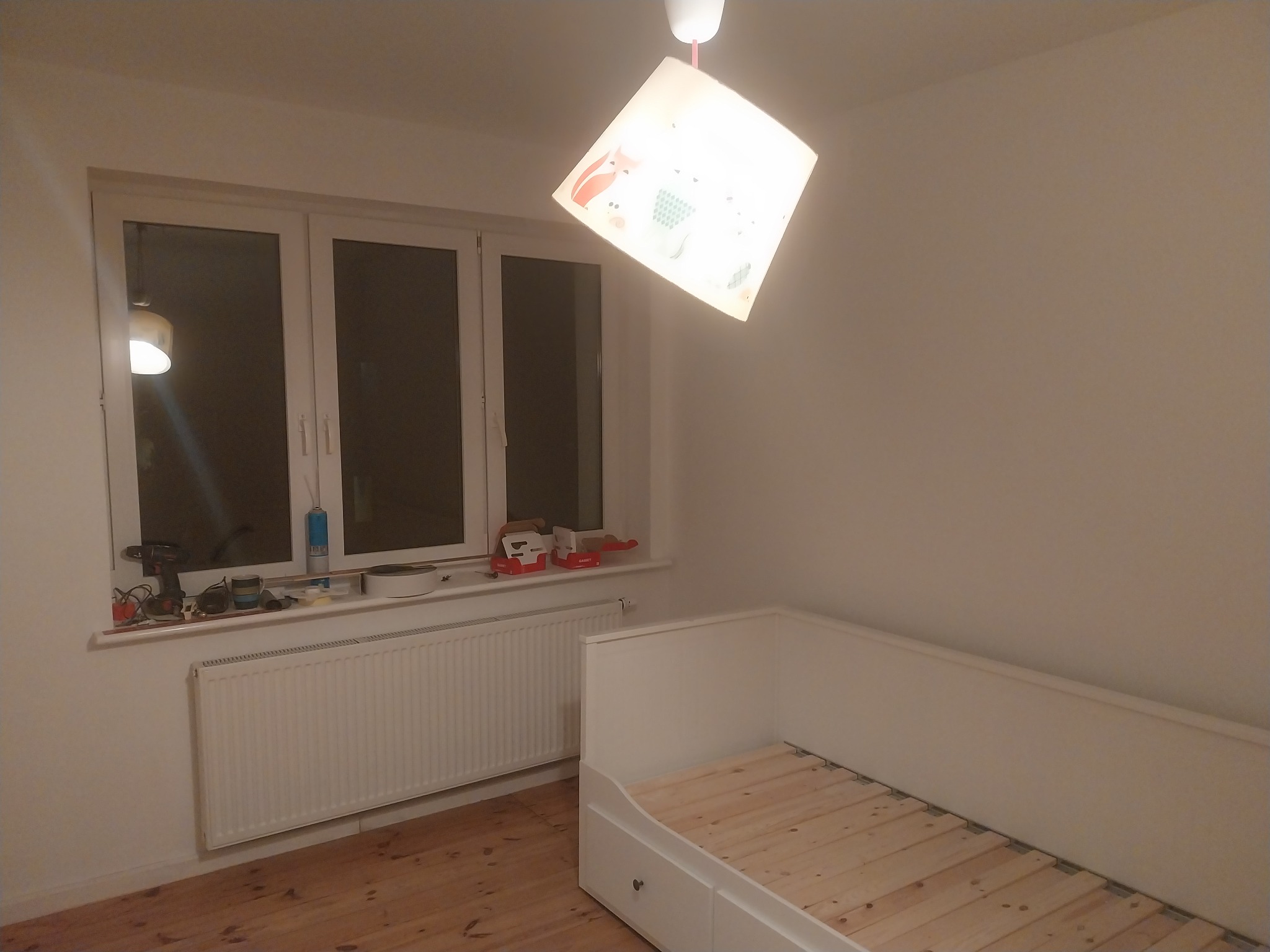 Pokój dziecięcy w trakcie malowania: białe ściany, drewniana podłoga, białe łóżko z szufladami, grzejnik pod oknem z narzędziami i akcesoriami malarskimi na parapecie, lampa z motywem zwierzęcym.