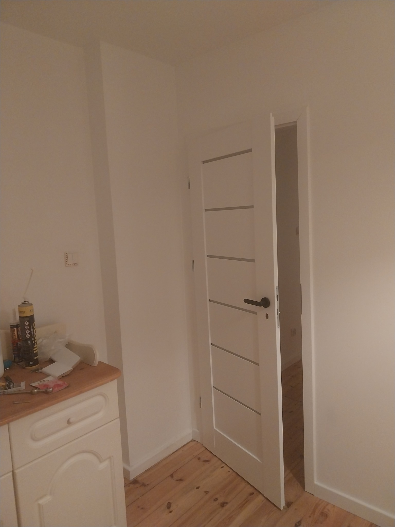 Świeżo pomalowane na biało ściany i drzwi wewnętrzne z poziomymi frezami, widoczne narzędzia malarskie na szafce i drewniana podłoga.