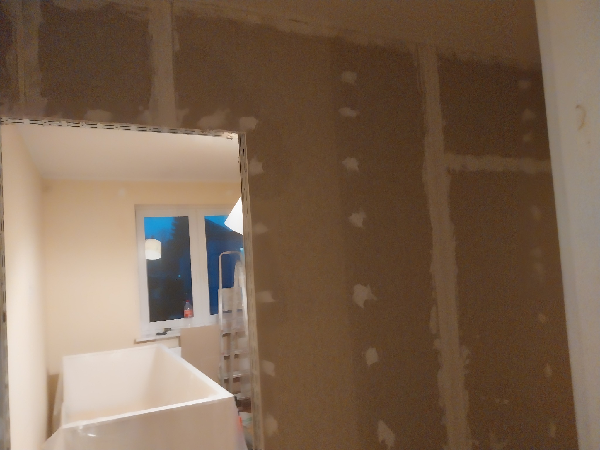 Ściana w trakcie szpachlowania, widoczne łączenia płyt gipsowo-kartonowych i punktowe uzupełnienia ubytków, w tle okno i drabina.