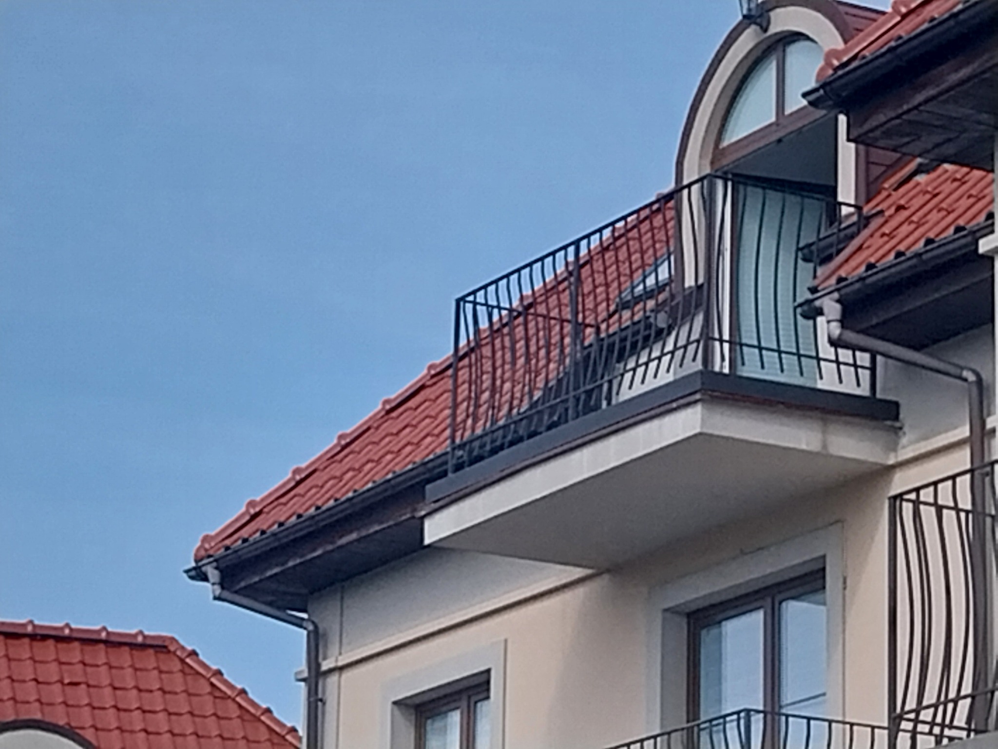 Fragment budynku z czerwonym dachem i balkonem z czarną, metalową balustradą, widok z dołu na tle błękitnego nieba.