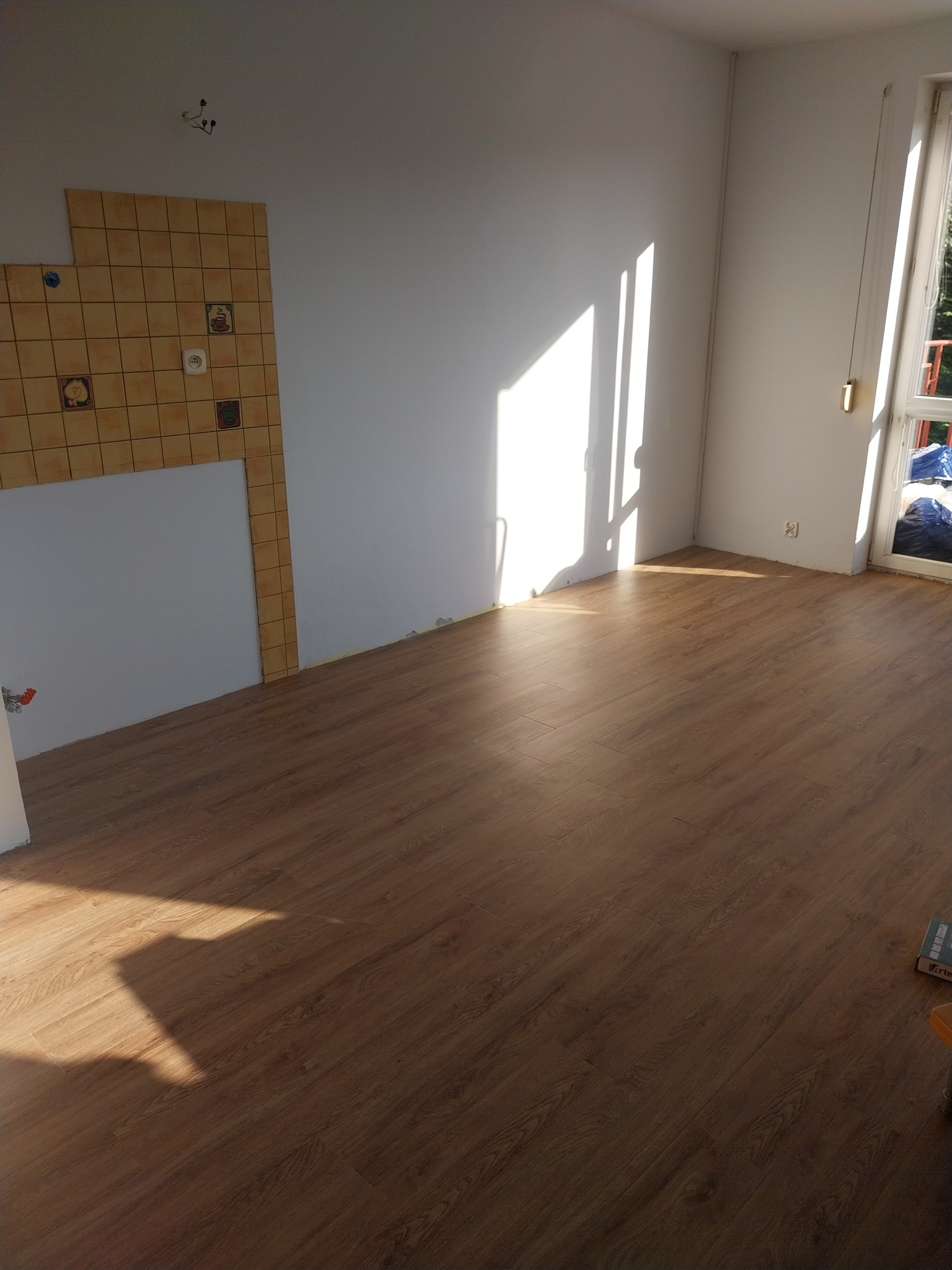 Pusty pokój po remoncie z nową, brązową podłogą, fragmentem ściany z niedokończonymi kafelkami i promieniami słońca wpadającymi przez okno.