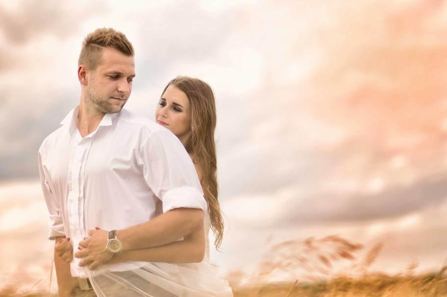 Para obejmująca się na tle zachodzącego słońca, mężczyzna w białej koszuli i zegarku, kobieta z długimi włosami spogląda w górę