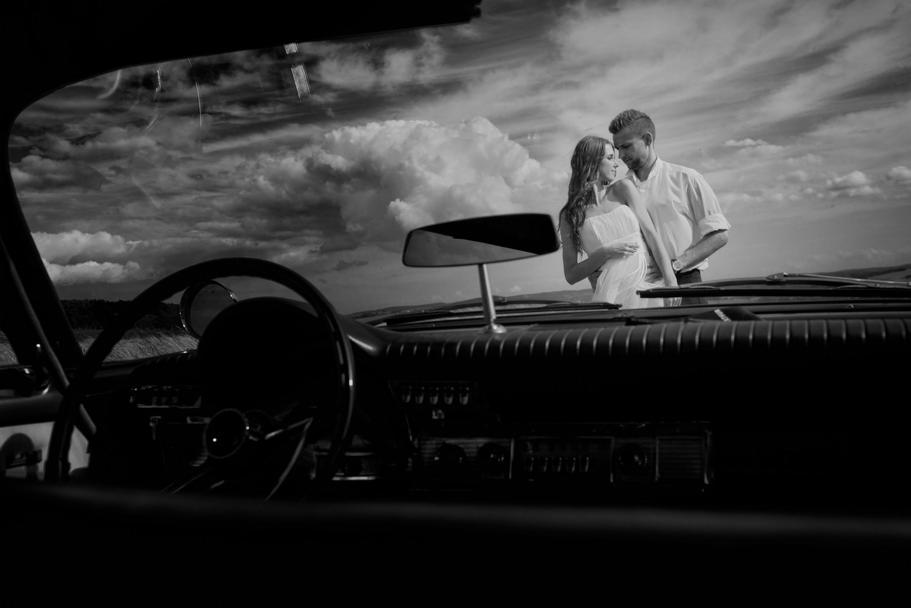 Czarno-białe zdjęcie pary narzeczeńskiej stojącej za klasycznym samochodem, widziane z wnętrza pojazdu, z dramatycznym tłem chmur.