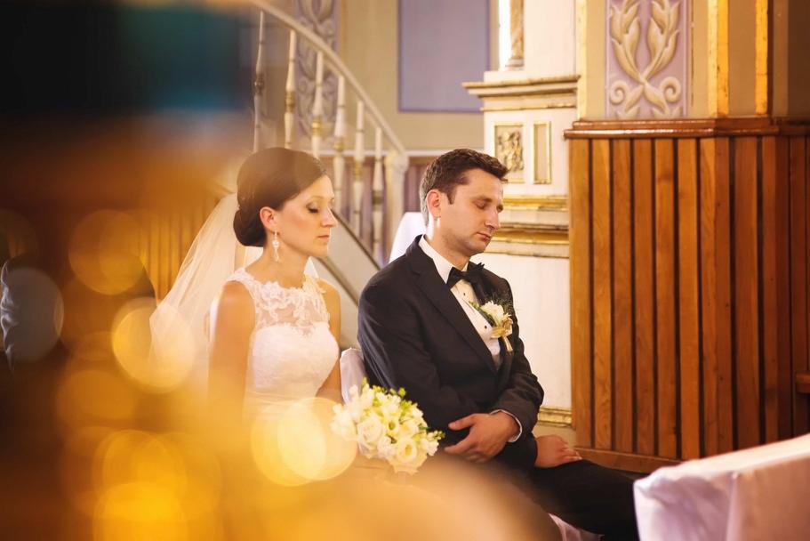 Para młoda siedzi w kościele podczas ceremonii ślubnej, panna młoda w koronkowej sukni i welonie, pan młody w czarnym smokingu, w tle drewniane panele i schody.