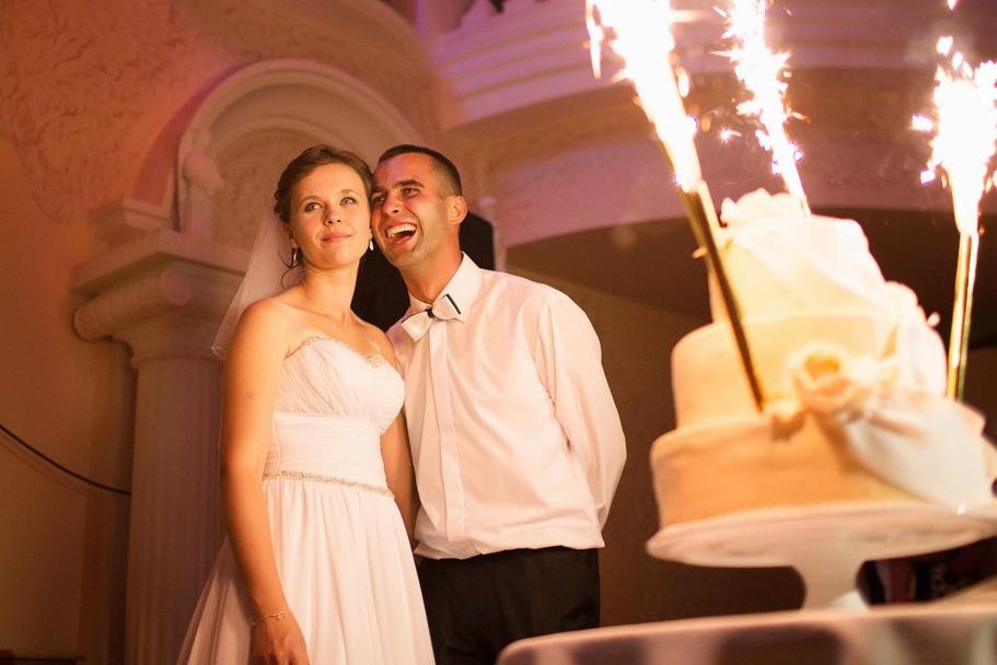 Uśmiechnięta para młoda w białych strojach, z tortem weselnym ozdobionym płonącymi racami, w tle kolumna i łuk.