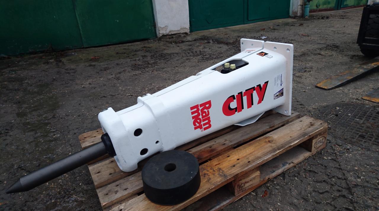 Biały młot hydrauliczny Rammer City na drewnianej palecie, z widocznym czarnym amortyzatorem i fragmentem wózka widłowego w tle.