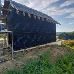 Budowa domu w trakcie: ściany pokryte czarną membraną z napisem DACHOWA, widoczne elementy więźby dachowej, rusztowanie. W tle zieleń i błękitne niebo.