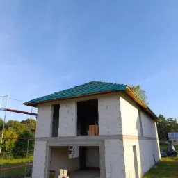 Niedokończony dwukondygnacyjny dom z białego bloczka, z zielonym dachem w trakcie budowy. Widoczne rusztowanie i materiały budowlane. Błękitne niebo w tle.