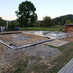 Przygotowany teren pod fundamenty domu z widoczną izolacją, podsypką żwirową i częściowo zalaną płytą fundamentową na tle zielonego krajobrazu.