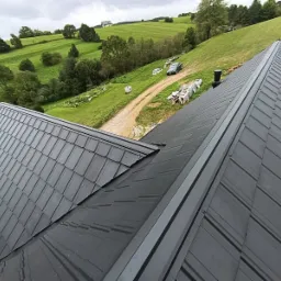 Czarny dach z blachy na tle zielonych wzgórz. Widoczny fragment konstrukcji dachu z czerwoną membraną. Ujęcie z góry, podkreślające geometrię pokrycia.