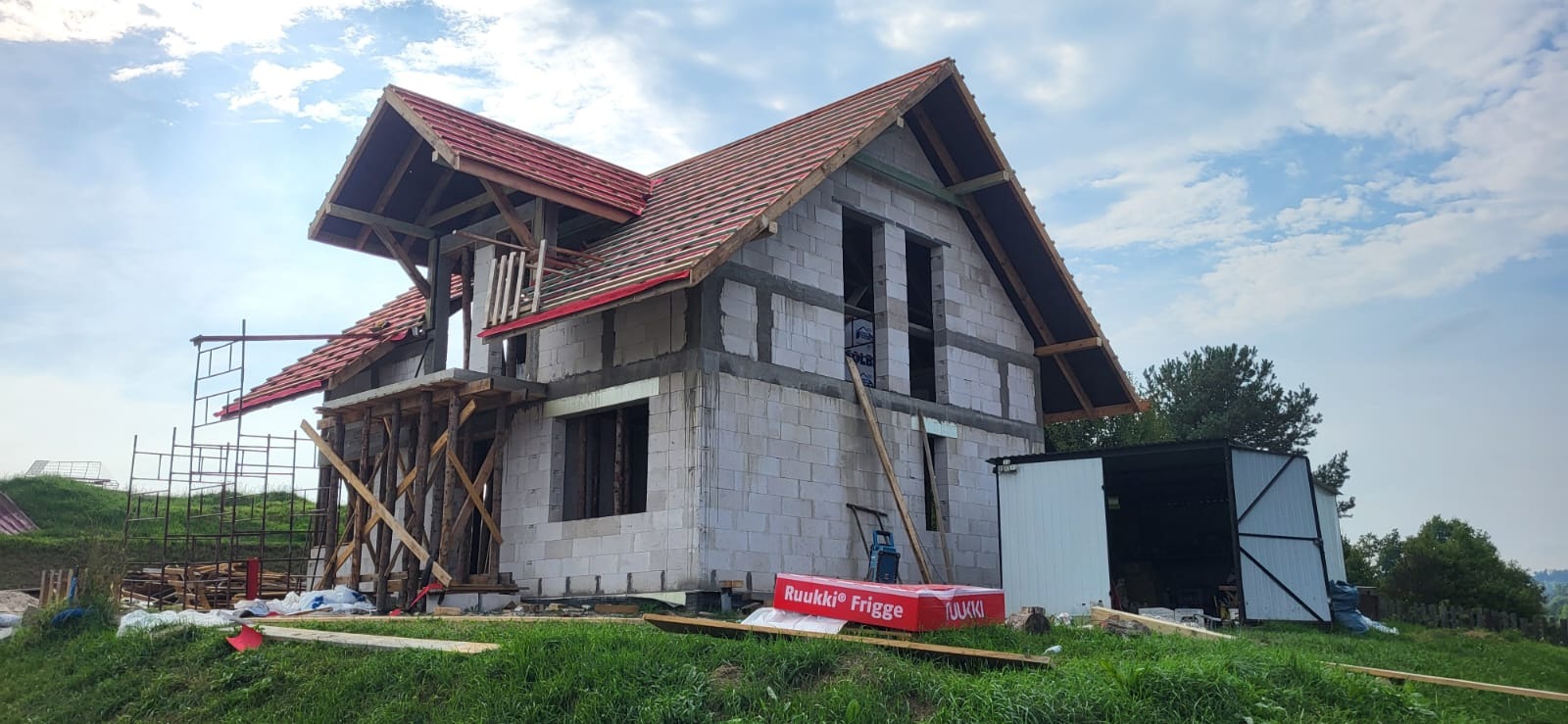 Budowa domu z białych bloczków, częściowo pokryty czerwoną dachówką. Widoczne drewniane rusztowania i materiały budowlane. Etap wznoszenia konstrukcji.