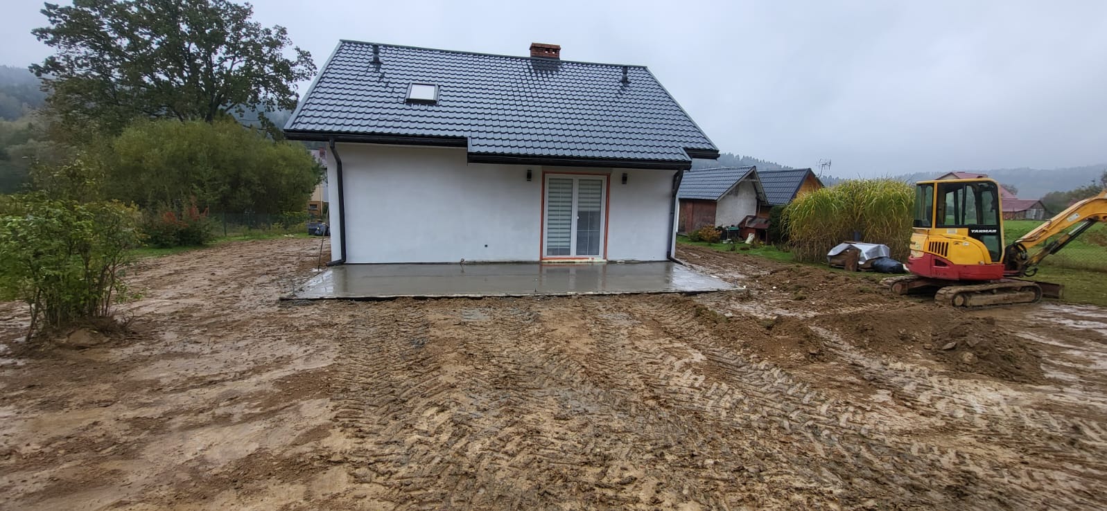 Nowoczesny dom z ciemnym dachem i świeżo wylanym betonowym tarasem w Ropience. Żółto-czerwona koparka na błotnistym terenie sugeruje prace ziemne.