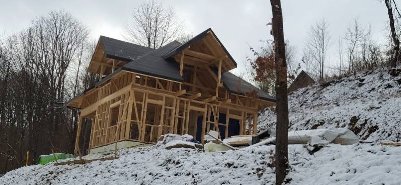 Dom szkieletowy w trakcie budowy, widoczna drewniana konstrukcja i fragment dachu na zasypanym śniegiem wzgórzu. Pochmurne niebo w tle.