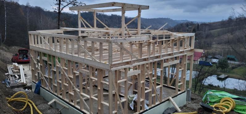 Surowy stan domu szkieletowego w trakcie budowy, widoczne drewniane ramy i fundamenty na tle pagórkowatego krajobrazu z jeziorem i lasem. W tle czerwony samochód.