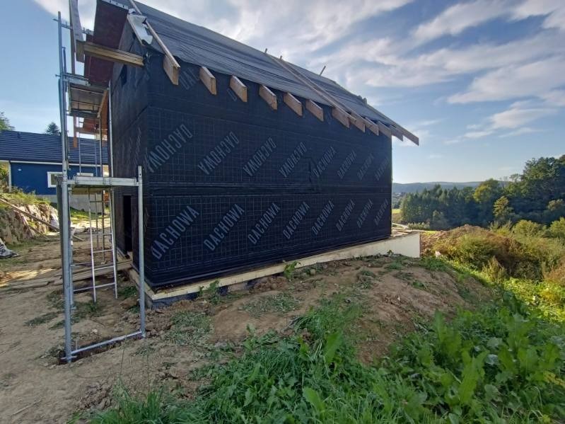 Budowa domu w trakcie: ściany pokryte czarną membraną z napisem DACHOWA, widoczne elementy więźby dachowej, rusztowanie. W tle zieleń i błękitne niebo.