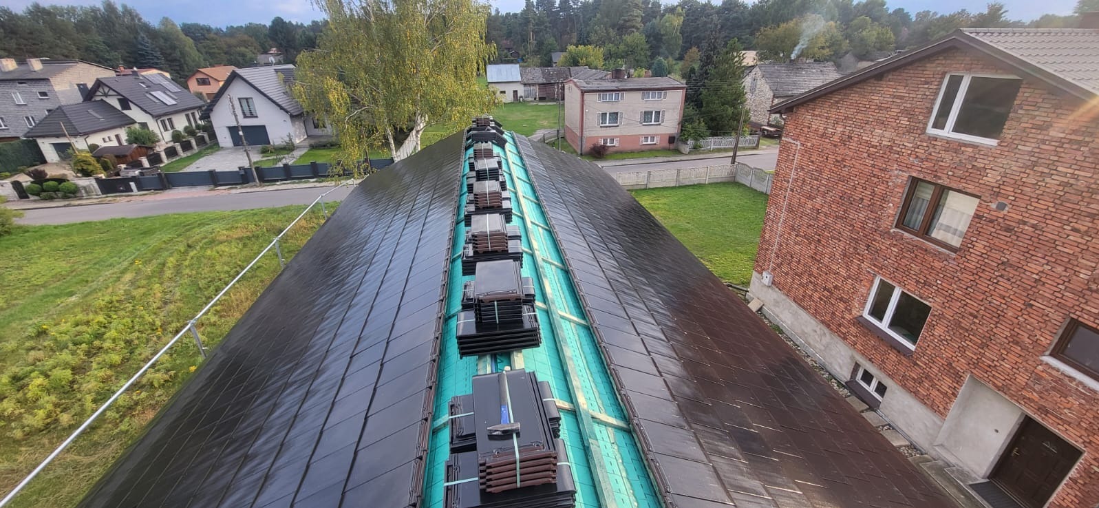 Układanie ciemnej dachówki na dachu domu. Widoczne przygotowane pakiety dachówek i fragmenty ułożonej połaci. W tle zabudowa jednorodzinna i zieleń.