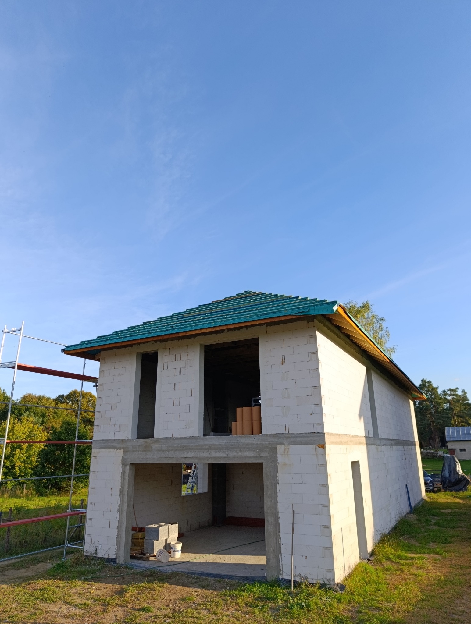 Niedokończony dwukondygnacyjny dom z białego bloczka, z zielonym dachem w trakcie budowy. Widoczne rusztowanie i materiały budowlane. Błękitne niebo w tle.