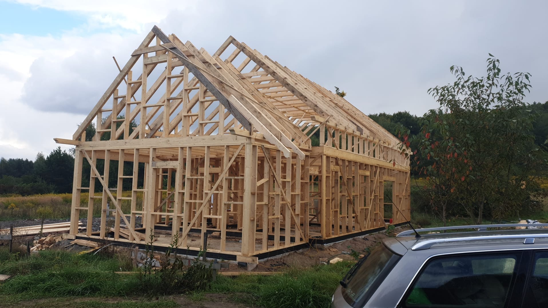 Konstrukcja domu szkieletowego w trakcie budowy, widoczne drewniane ramy ścian i dachu na tle zieleni i pochmurnego nieba. Fragment samochodu w prawym dolnym rogu.
