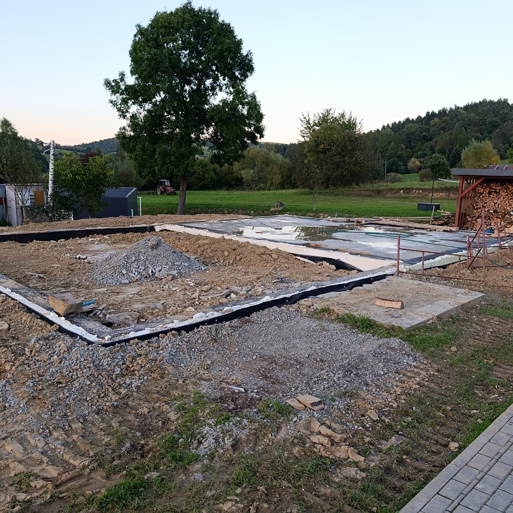 Przygotowany teren pod fundamenty domu z widoczną izolacją, podsypką żwirową i częściowo zalaną płytą fundamentową na tle zielonego krajobrazu.