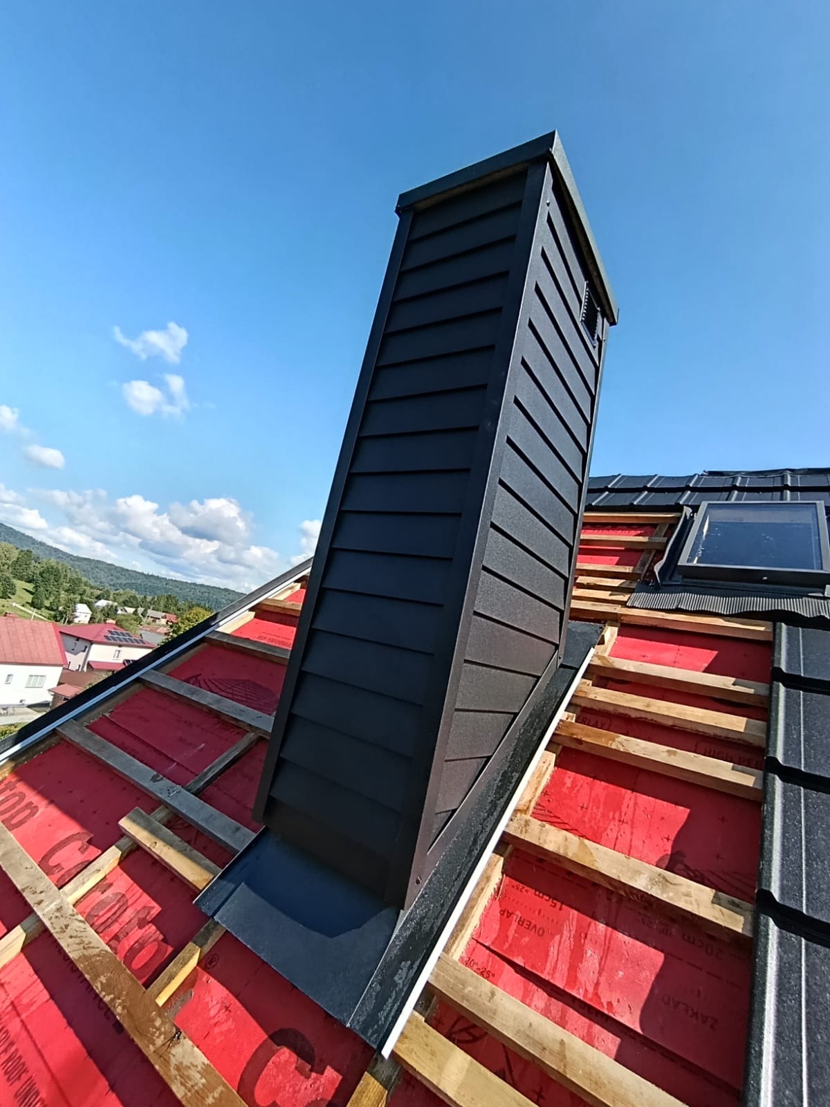 Montaż obudowy komina z blachy na dachu z oknem połaciowym. Widoczne łaty, membrana dachowa i fragmenty krajobrazu w tle. Kątowe ujęcie z dołu.