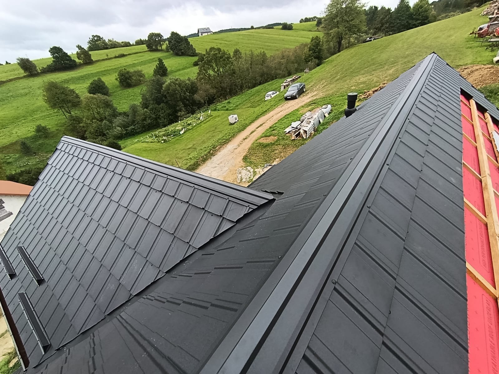 Czarny dach z blachy na tle zielonych wzgórz. Widoczny fragment konstrukcji dachu z czerwoną membraną. Ujęcie z góry, podkreślające geometrię pokrycia.
