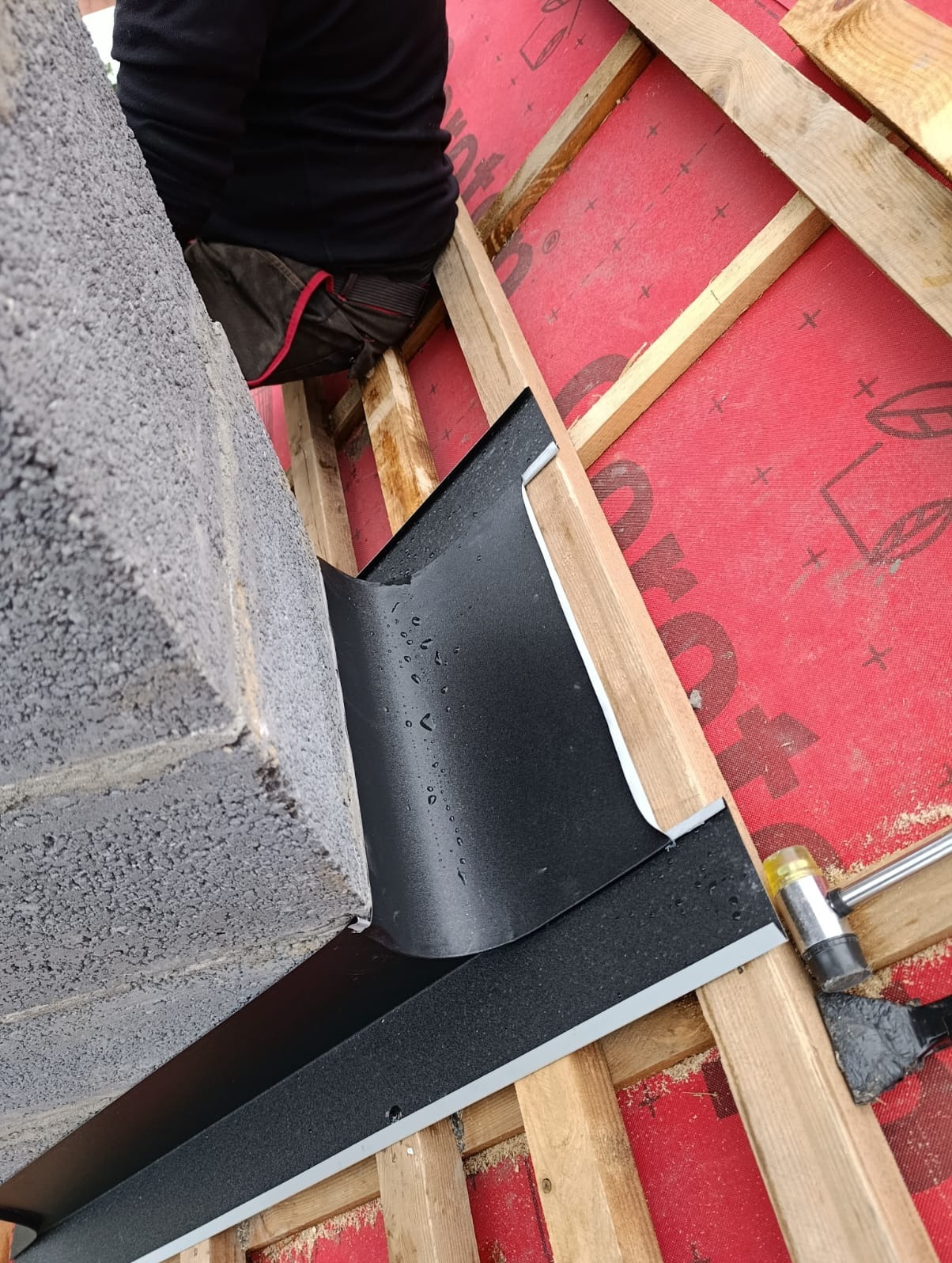 Montaż obróbki blacharskiej komina na dachu z membraną, widoczne drewniane elementy konstrukcji dachu i narzędzia. Obróbka w kolorze czarnym, lekko wilgotna.