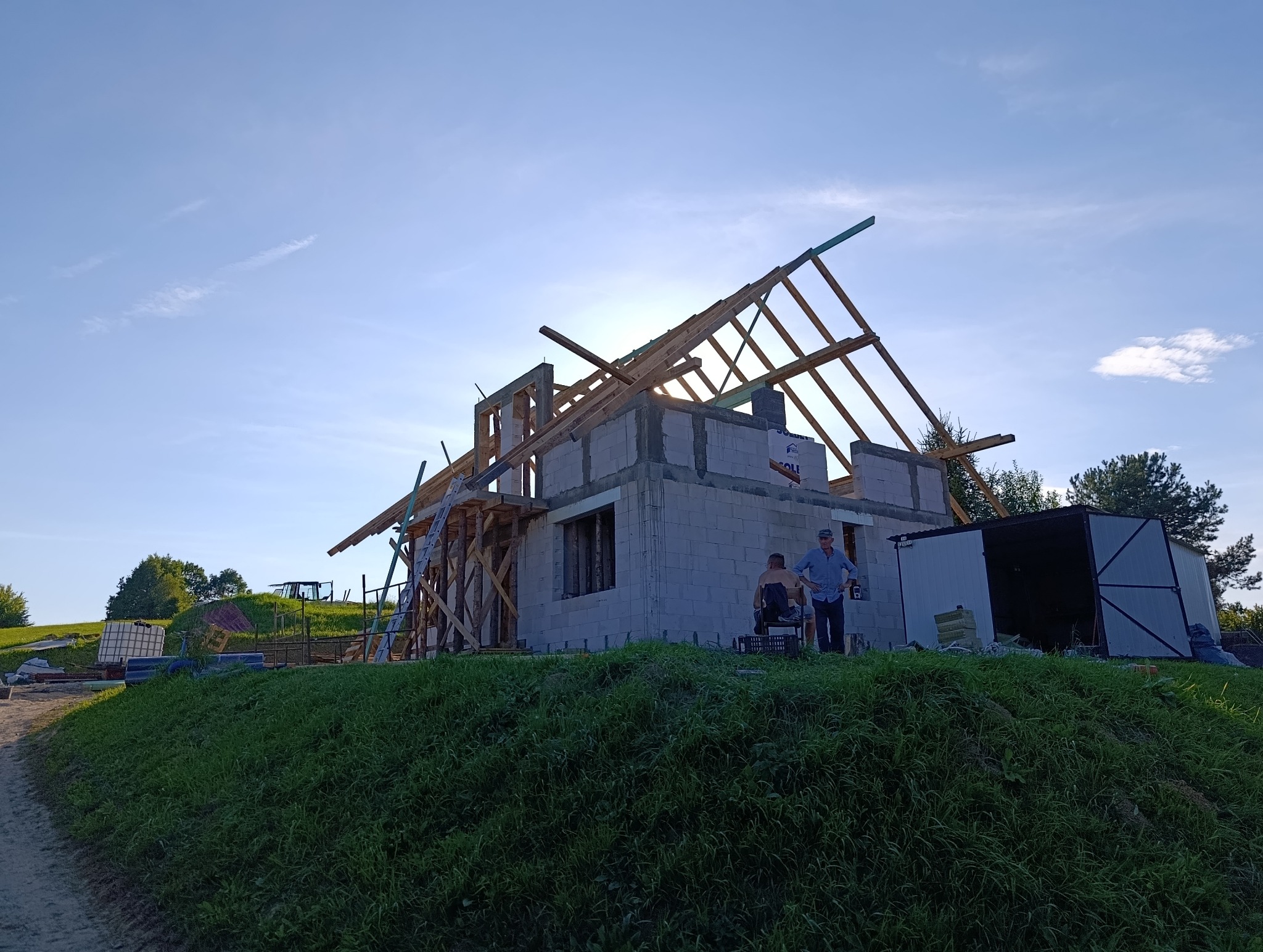 Budowa domu z betonu komórkowego w trakcie wznoszenia więźby dachowej. Dwóch mężczyzn przy ścianie budynku. Widok z dołu na tle błękitnego nieba.