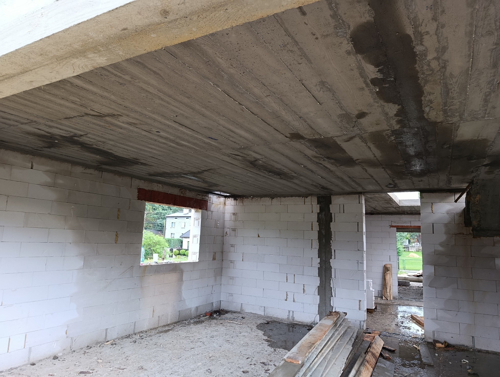 Surowy stan domu z betonu komórkowego: ściany z pustaków, betonowy strop z widocznymi śladami szalunków, okno z widokiem na zieleń. Wnętrze w trakcie budowy.