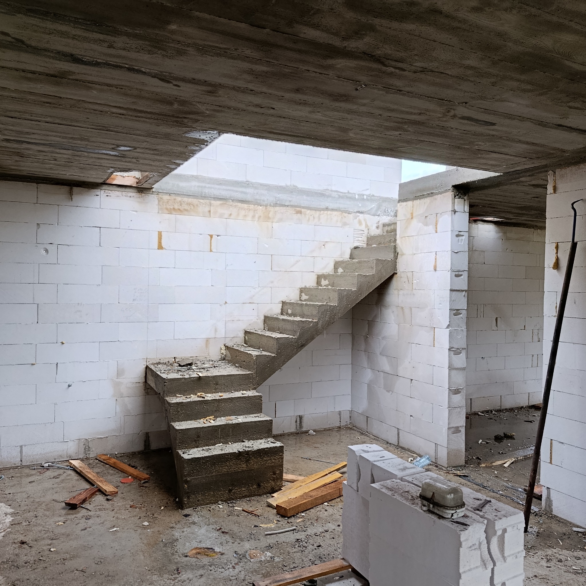 Surowe betonowe schody wewnątrz budynku z białych bloczków, widoczne niedokończone elementy konstrukcyjne i narzędzia budowlane. W tle otwór w stropie.