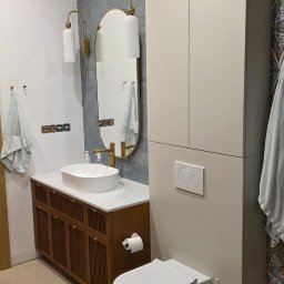 Magic Meble - Nowoczesna łazienka z szafką pod umywalką z drewnianymi lamelami, owalnym lustrem w złotej ramie, i zabudową nad toaletą w odcieniach beżu.