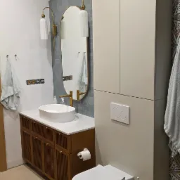 Nowoczesna łazienka z szafką pod umywalką z drewnianymi lamelami, owalnym lustrem w złotej ramie, i zabudową nad toaletą w odcieniach beżu.