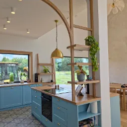 Błękitna kuchnia z drewnianym blatem i dekoracyjną ścianką działową z półkami. Widok na zewnątrz przez okno z ciężarówką. Wnętrze w stylu nowoczesnym z elementami rustykalnymi.