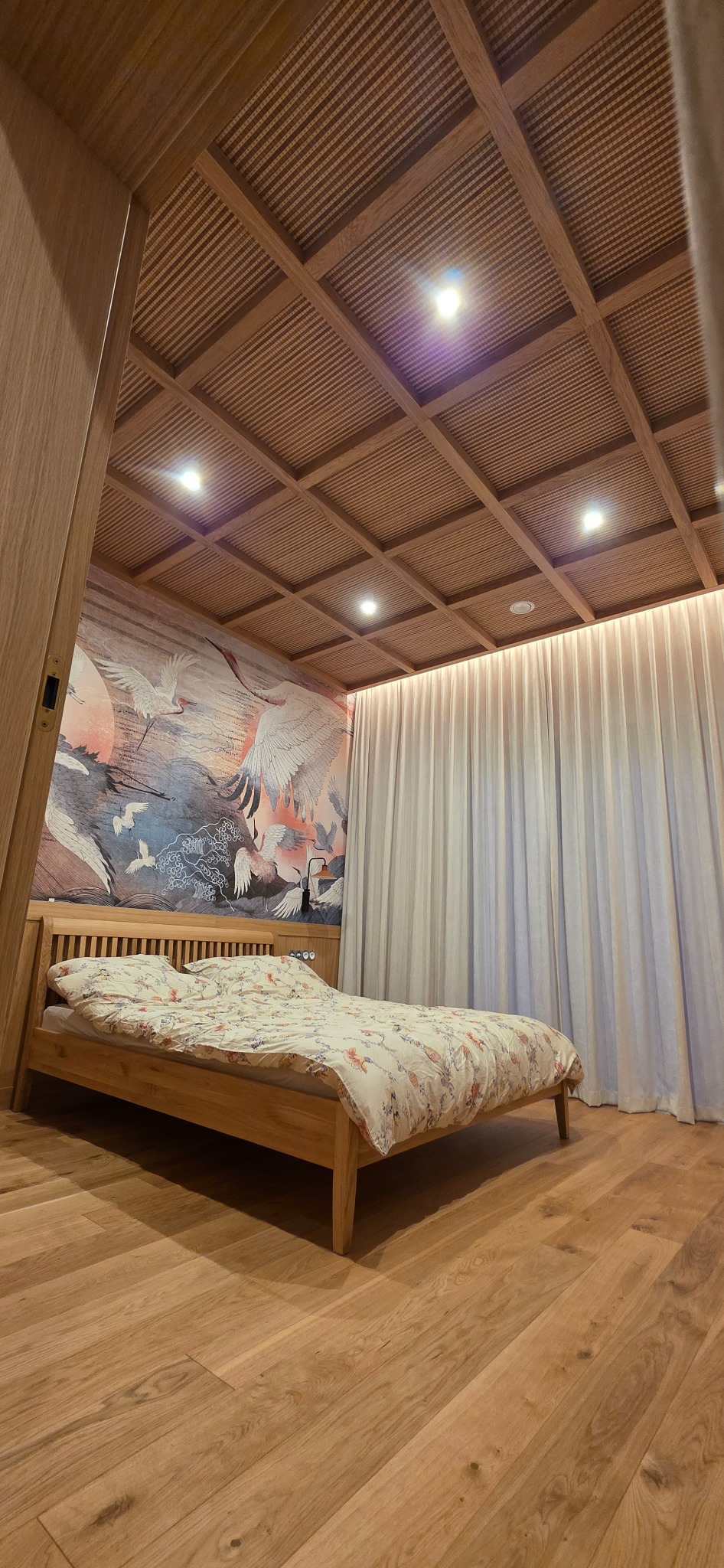 Drewniane wykończenie sypialni: parkiet, łóżko z zagłówkiem, sufit kasetonowy z oświetleniem. Na ścianie fototapeta z motywem żurawi. Lekkie, zwiewne zasłony.
