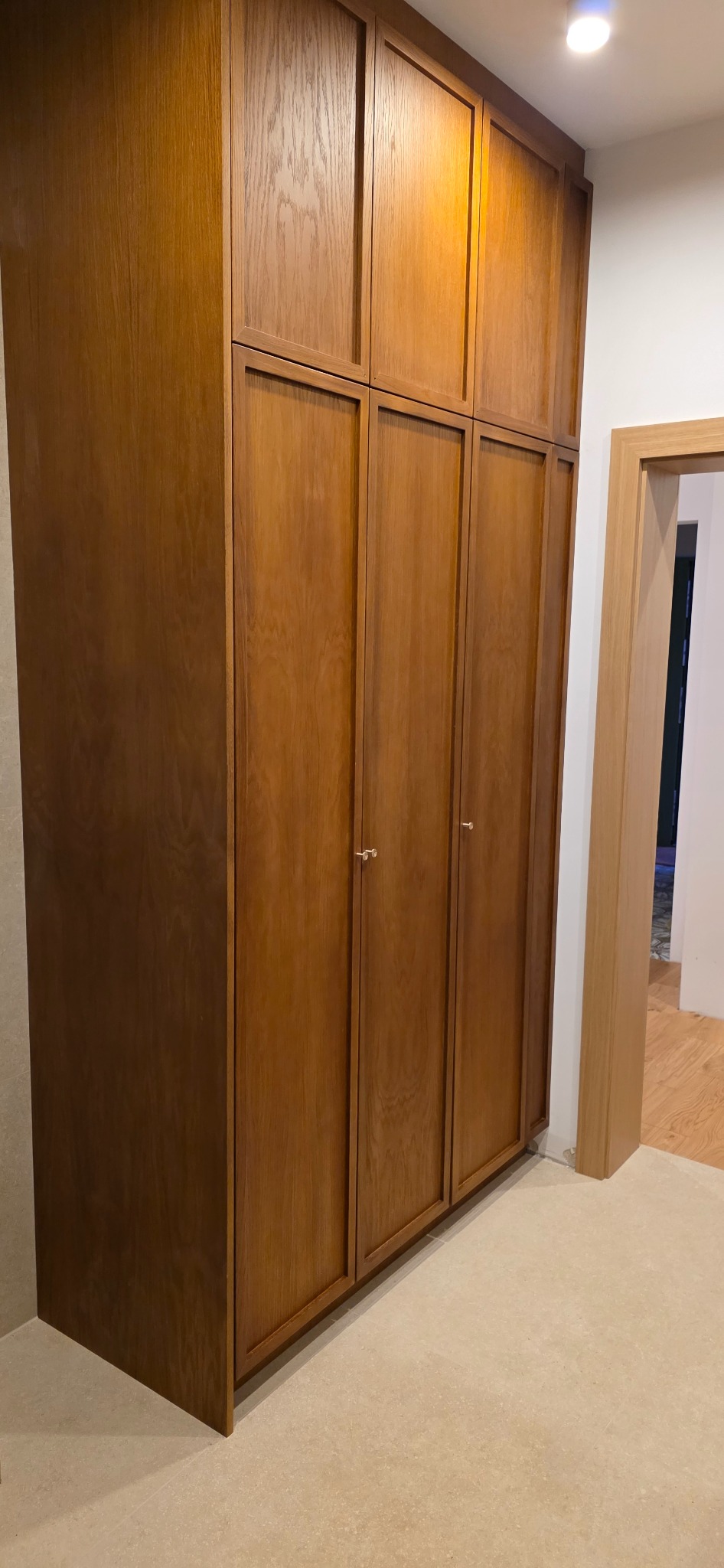 Drewniana szafa wnękowa z czterema drzwiami i górnymi szafkami, z minimalistycznymi, okrągłymi uchwytami, z widocznym fragmentem drzwi po prawej stronie.