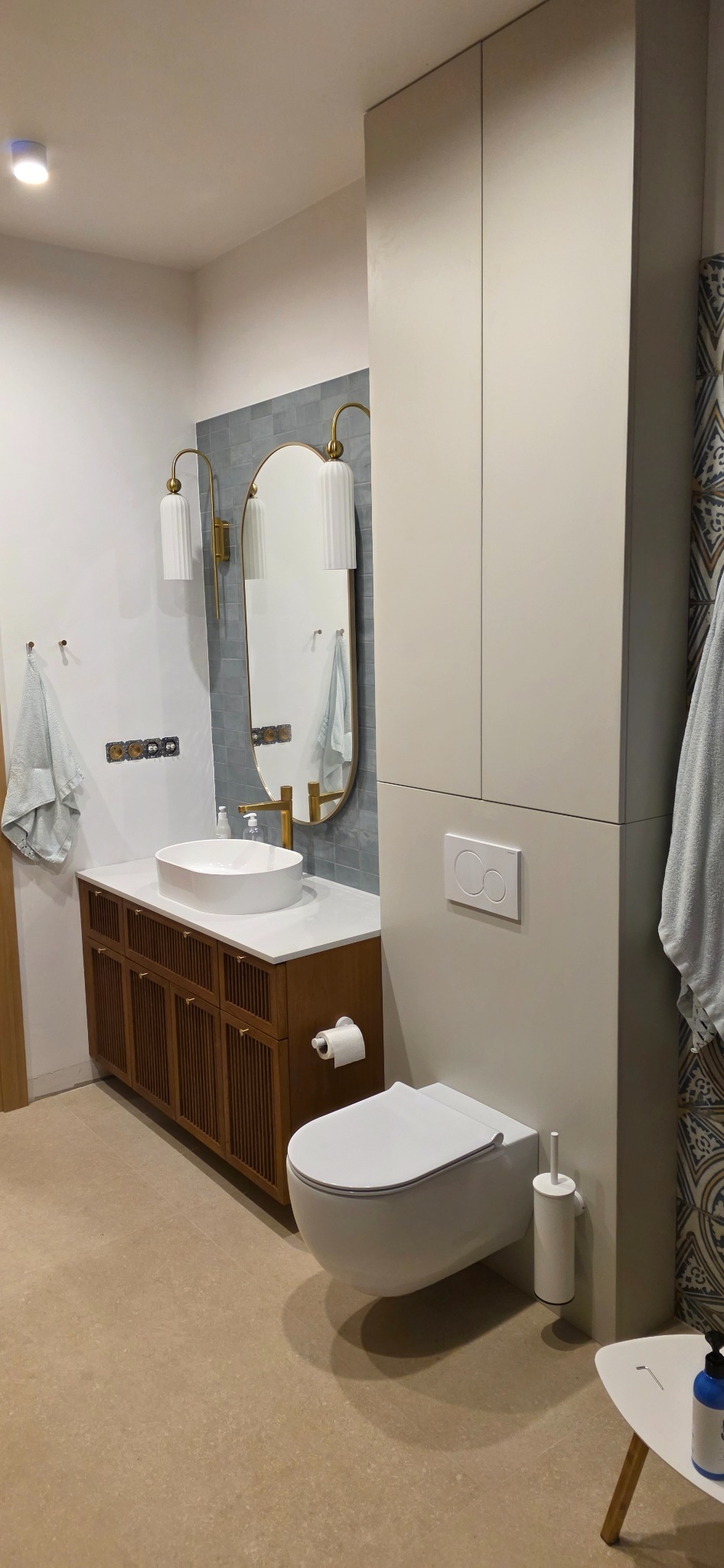 Nowoczesna łazienka z szafką pod umywalką z drewnianymi lamelami, owalnym lustrem w złotej ramie, i zabudową nad toaletą w odcieniach beżu.