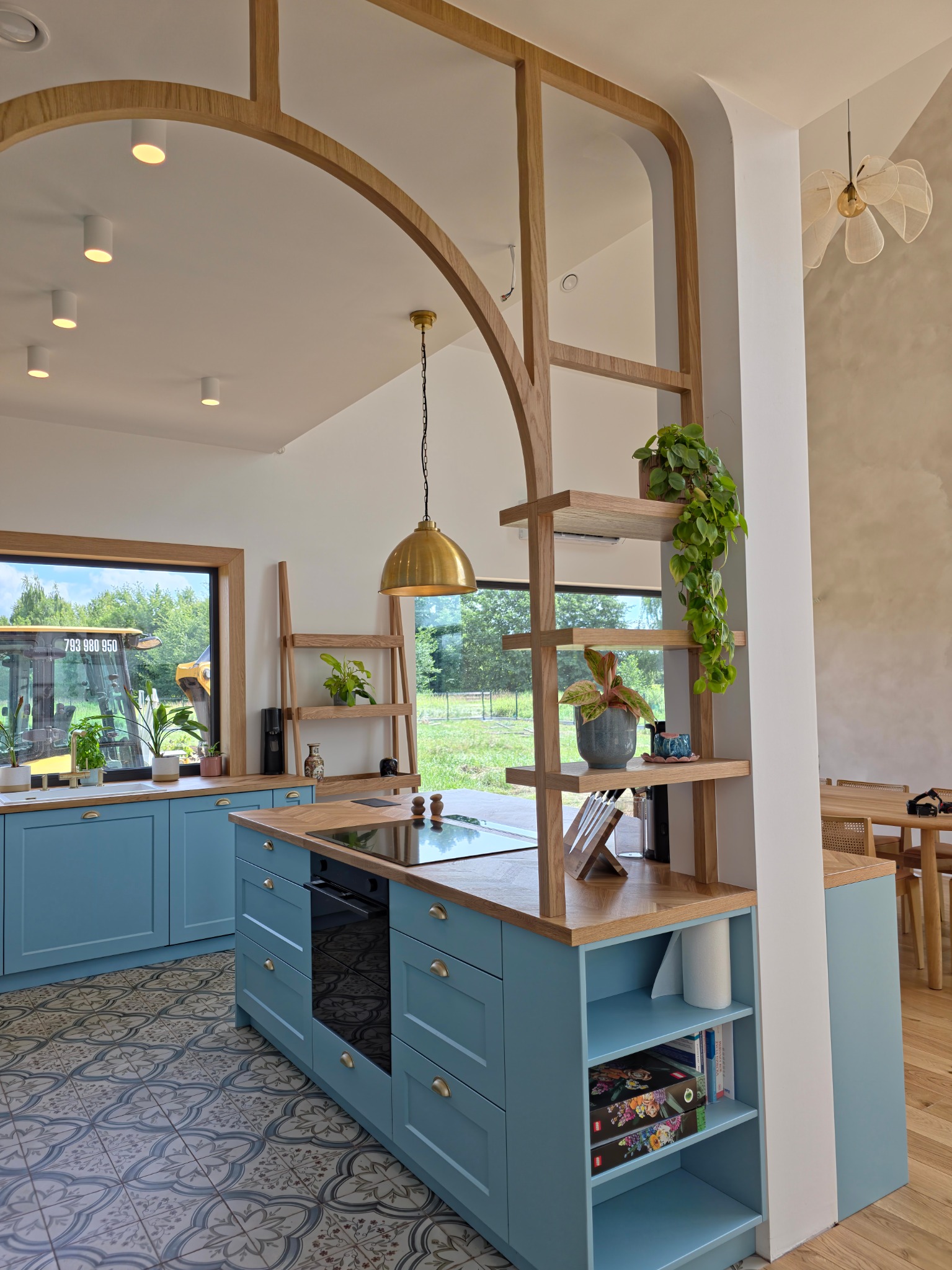 Błękitna kuchnia z drewnianym blatem i dekoracyjną ścianką działową z półkami. Widok na zewnątrz przez okno z ciężarówką. Wnętrze w stylu nowoczesnym z elementami rustykalnymi.