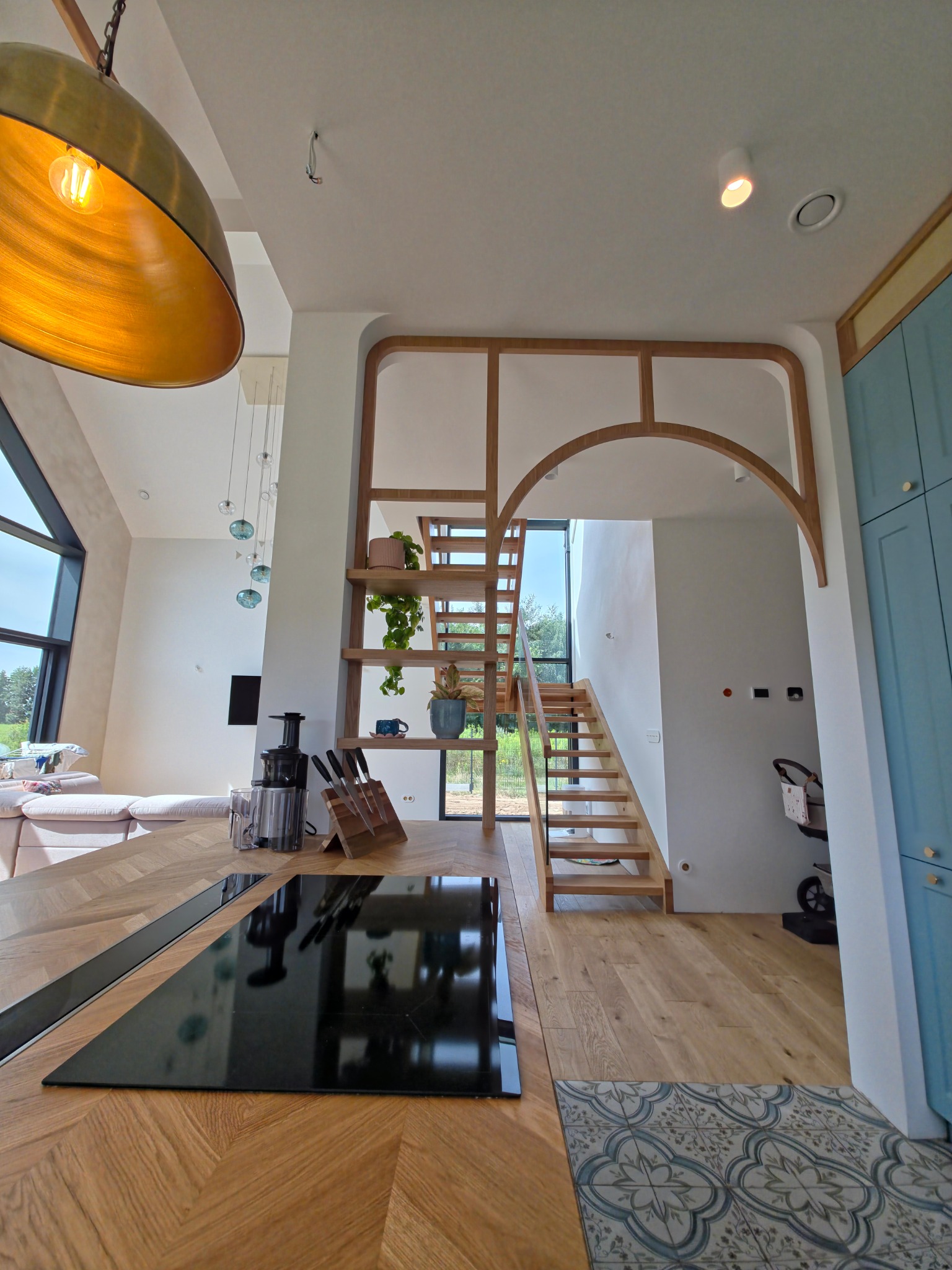 Nowoczesna kuchnia z drewnianą wyspą i płytą indukcyjną, drewniane schody z półkami na rośliny, minimalistyczne oświetlenie i błękitne szafki.