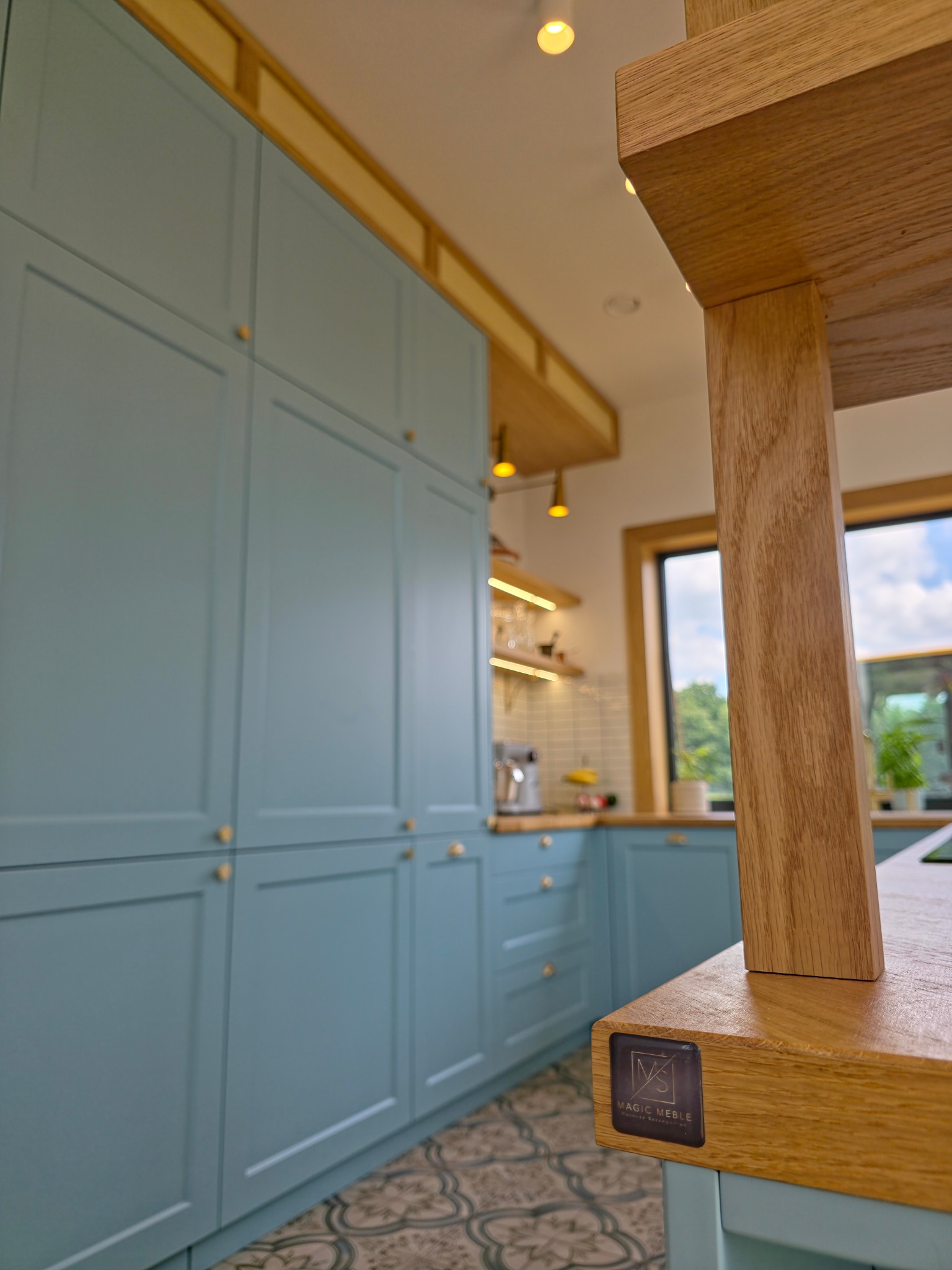 Błękitna kuchnia na wymiar z drewnianym blatem w Twardogórze. Klasyczne fronty, złote gałki, wzorzysta podłoga. Widoczny fragment okna i oświetlenia.