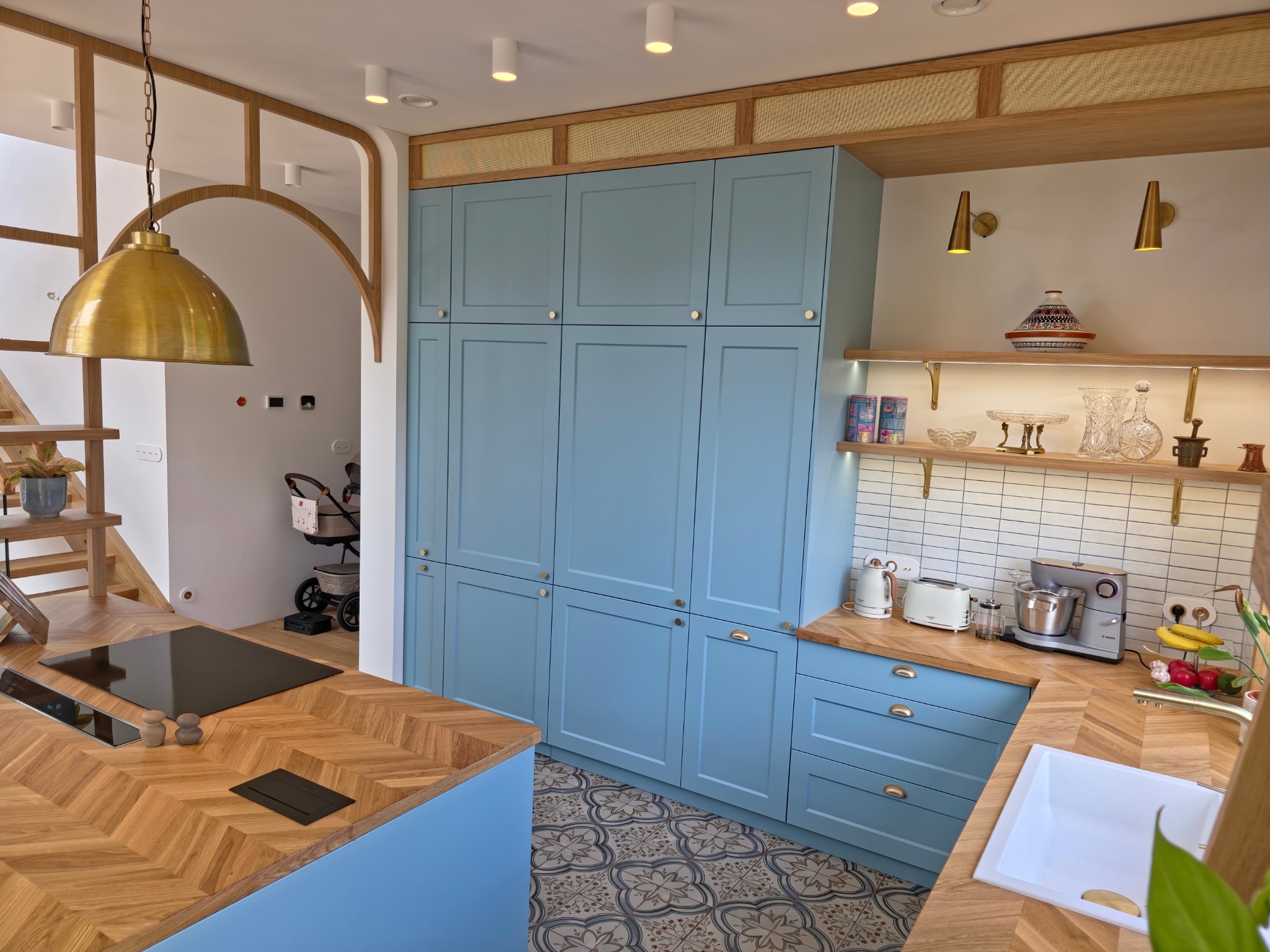 Niebieska zabudowa kuchenna z drewnianym blatem w jodełkę i złotymi akcentami. Widoczna wyspa kuchenna i elementy dekoracyjne na półkach.