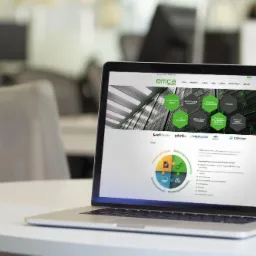 Laptop wyświetlający stronę internetową IT EMCA na białym stole w biurze, z widocznymi elementami graficznymi i logo.