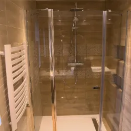 Nowoczesna kabina prysznicowa z chromowanymi profilami i deszczownicą, jasne płytki ceramiczne z dekoracyjnym pasem, biały grzejnik drabinkowy po lewej stronie.