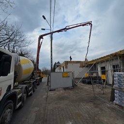 DKK PROPERTY INVESTMENTS SPÓŁKA Z OGRANICZONĄ ODPOWIEDZIALNOŚCIĄ - Betoniarka i pompa do betonu na budowie domu z białych bloczków. Widoczni pracownicy na dachu. Szare niebo w tle.