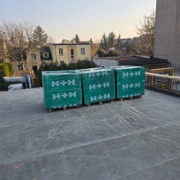 DKK PROPERTY INVESTMENTS SPÓŁKA Z OGRANICZONĄ ODPOWIEDZIALNOŚCIĄ - Silikatowe bloczki H+H na betonowym stropie budynku w trakcie budowy, widoczne zbrojenia i fragmenty sąsiednich budynków w tle, wczesne popołudnie.