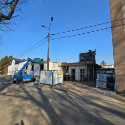 DKK PROPERTY INVESTMENTS SPÓŁKA Z OGRANICZONĄ ODPOWIEDZIALNOŚCIĄ - Budowa domu w toku: robotnicy na dachu, szalunki, materiały budowlane, podnośnik. Widok z poziomu gruntu, cień fotografa w kadrze. Słoneczny dzień.