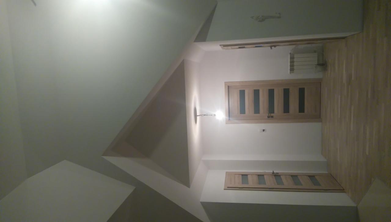 Wykończone wnętrze z drewnianą podłogą, białymi ścianami, dwiema drewnianymi ościeżnicami z szybami i oryginalnym sufitem z wnęką, oświetlone pojedynczą żarówką.