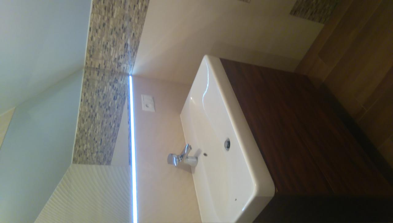 Umywalka w łazience z szafką w kolorze ciemnego drewna, mozaika na ścianie, podświetlenie LED i płytki imitujące drewno na podłodze.