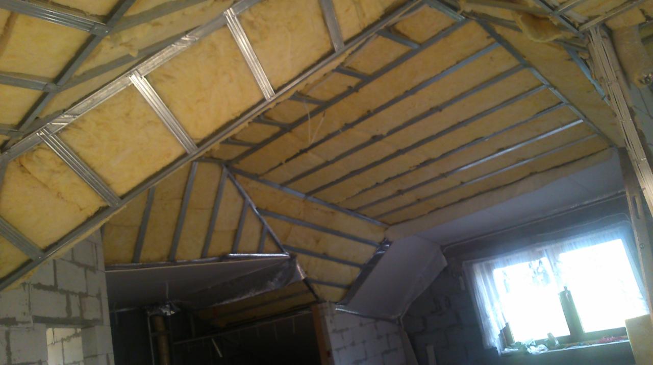 Montaż izolacji z wełny mineralnej w metalowym stelażu na poddaszu, widoczne ściany z bloczków betonowych i okno z białą firanką.