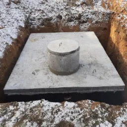 Betonowa płyta z włazem w wykopie ziemnym, widoczne metalowe uchwyty, lekko ośnieżona ziemia wokół.