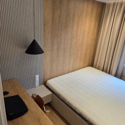 GLAZERIS Banakiewicz Adrian - Nowoczesna sypialnia po remoncie: drewniane panele ścienne, minimalistyczna lampa wisząca, biurko z laptopem i wygodne łóżko z materacem. Przytulne wnętrze.