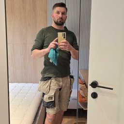 GLAZERIS Banakiewicz Adrian - Mężczyzna w odzieży roboczej robi selfie w lustrze na tle wykończonego wnętrza z łóżkiem i panelami ściennymi. Widoczne narzędzia w kieszeniach.